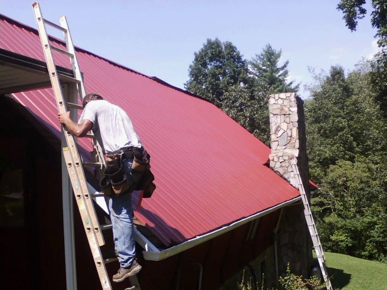 Roofer on a ladder beside a red metal roof and stone chimney — Soffit & Fascia Repair by Roofing Brothers in Phil Campbell